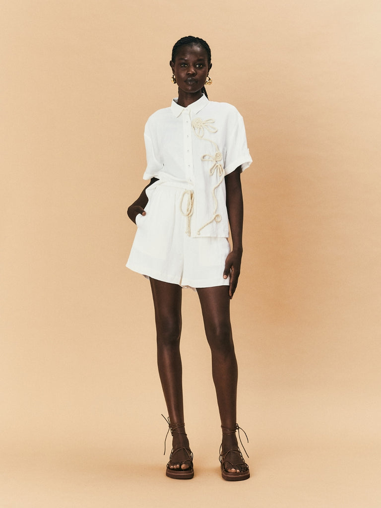 A young woman wearing La Maise shirt with white flower embroidery and La Maise Linen Shorts.