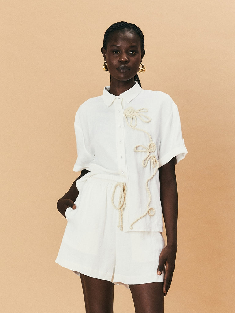 A young woman wearing La Maise Lillie shirt with white flower embroidery and La Maise linen shorts.