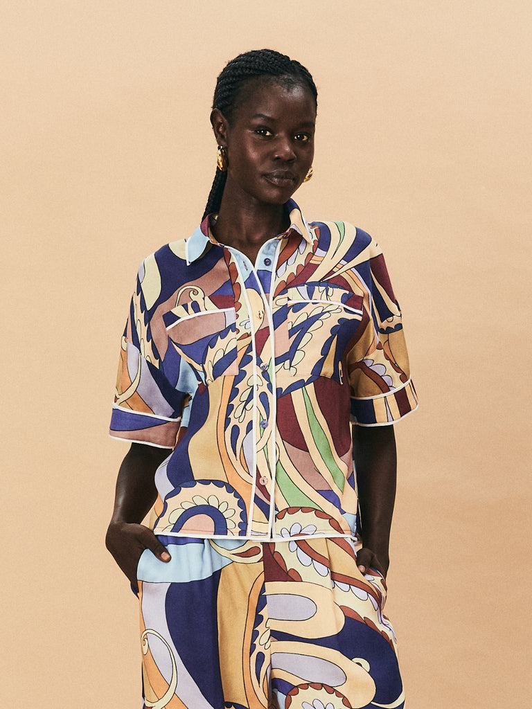 A young woman wearing a colourful patterned La Maise Dijon shirt against a beige background.