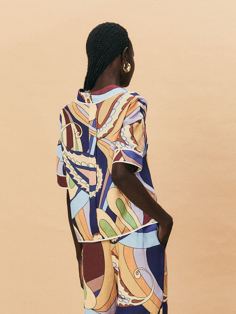 A young woman wearing a colourful patterned La Maise Dijon shirt against a beige background.
