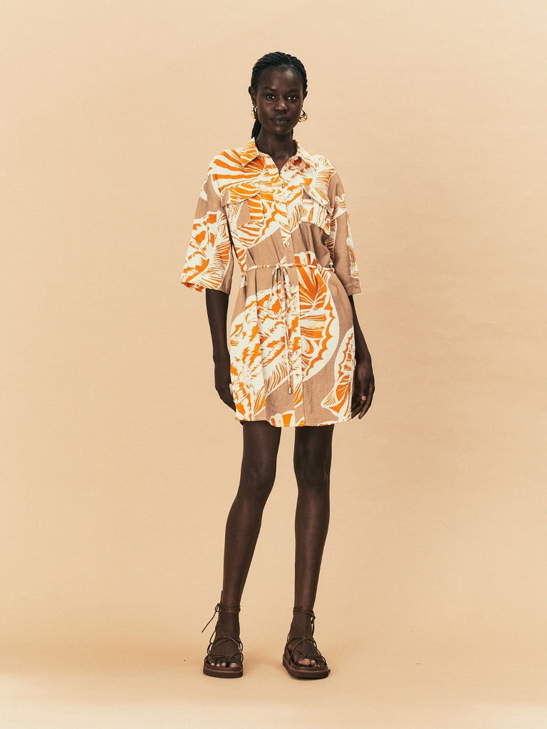 Woman wearing a patterned seashell print La Maise Cannes dress standing against a beige background.