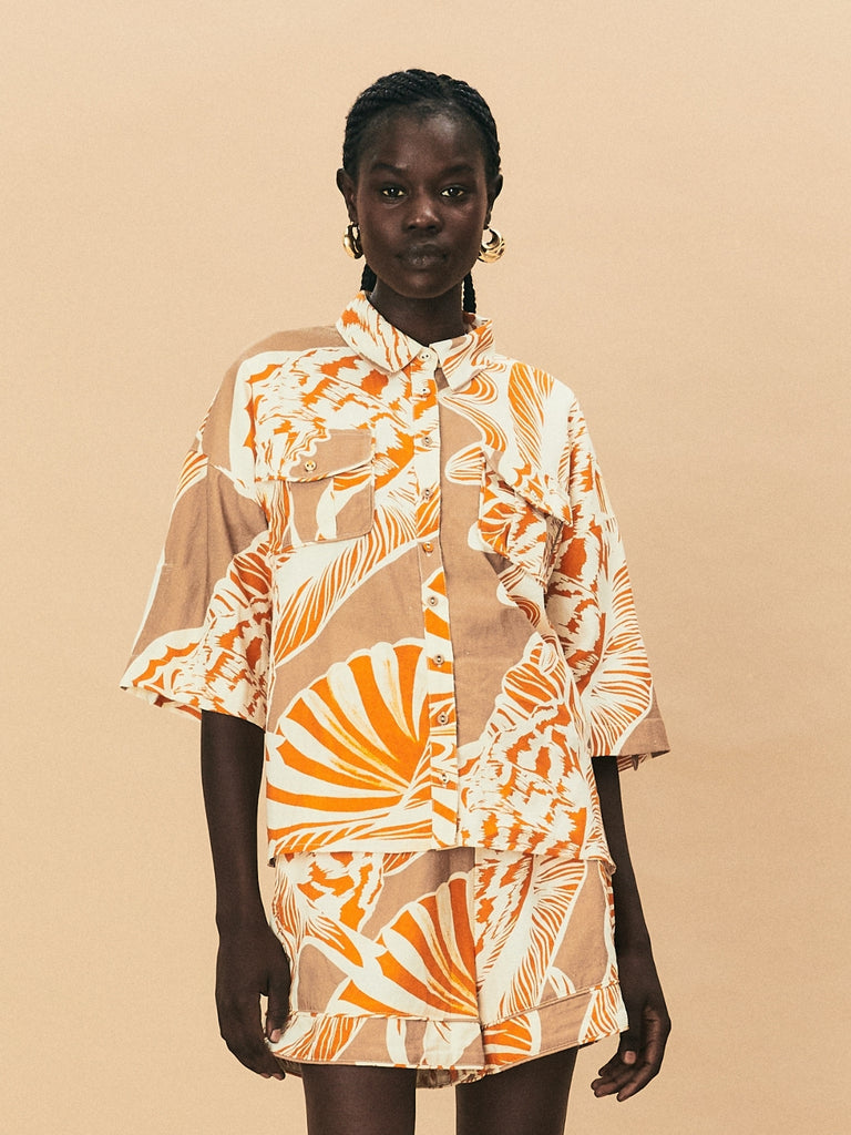 A young woman wearing La Maise Cannes linen shirt and shorts in seashell print against a beige background.