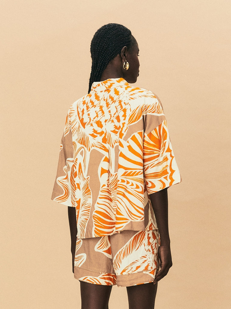 A young woman wearing La Maise Cannes linen shirt and shorts in seashell print against a beige background.