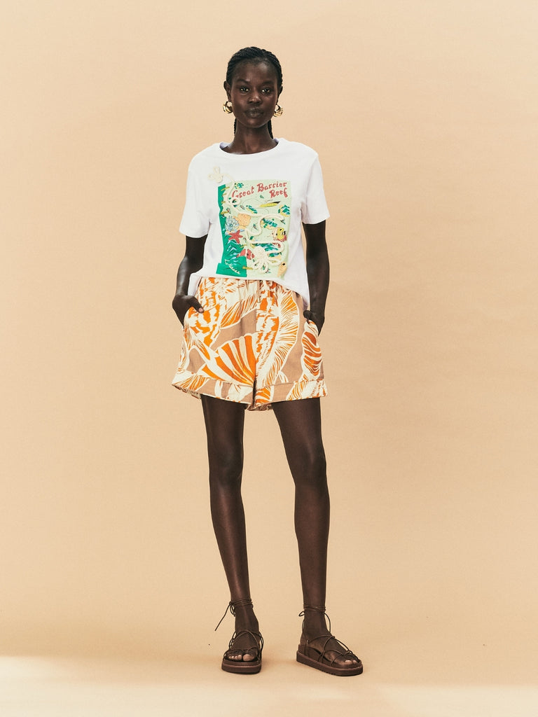 A young woman wearing a seashell print La Maise Cannes shorts with a white printed tee against a beige background.