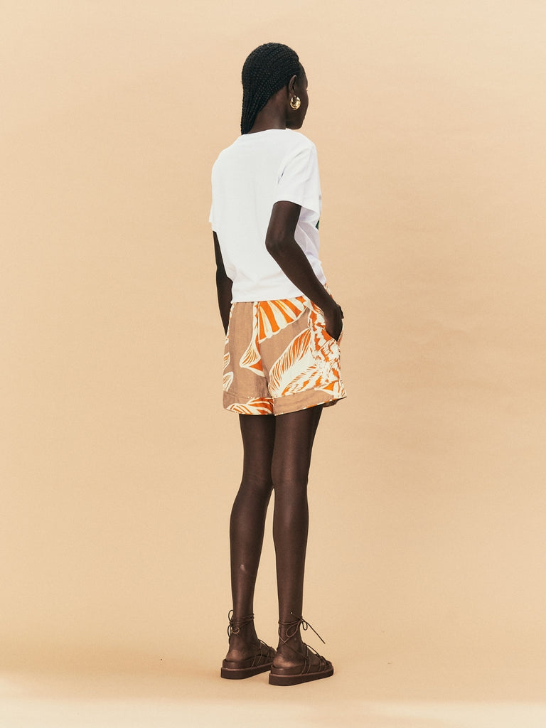 A young woman wearing a seashell print La Maise Cannes shorts with a white printed tee against a beige background.