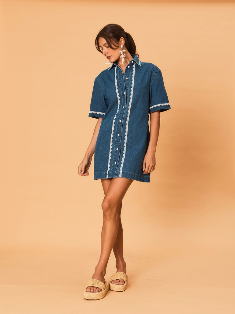A young woman wearing a blue La Maise Ellie denim dress with white lace trim on a beige background.