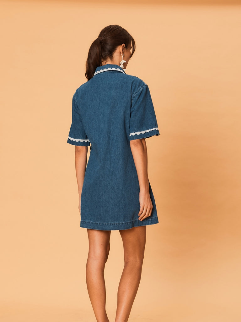 A young woman wearing a blue La Maise Ellie denim dress with white lace trim on a beige background.