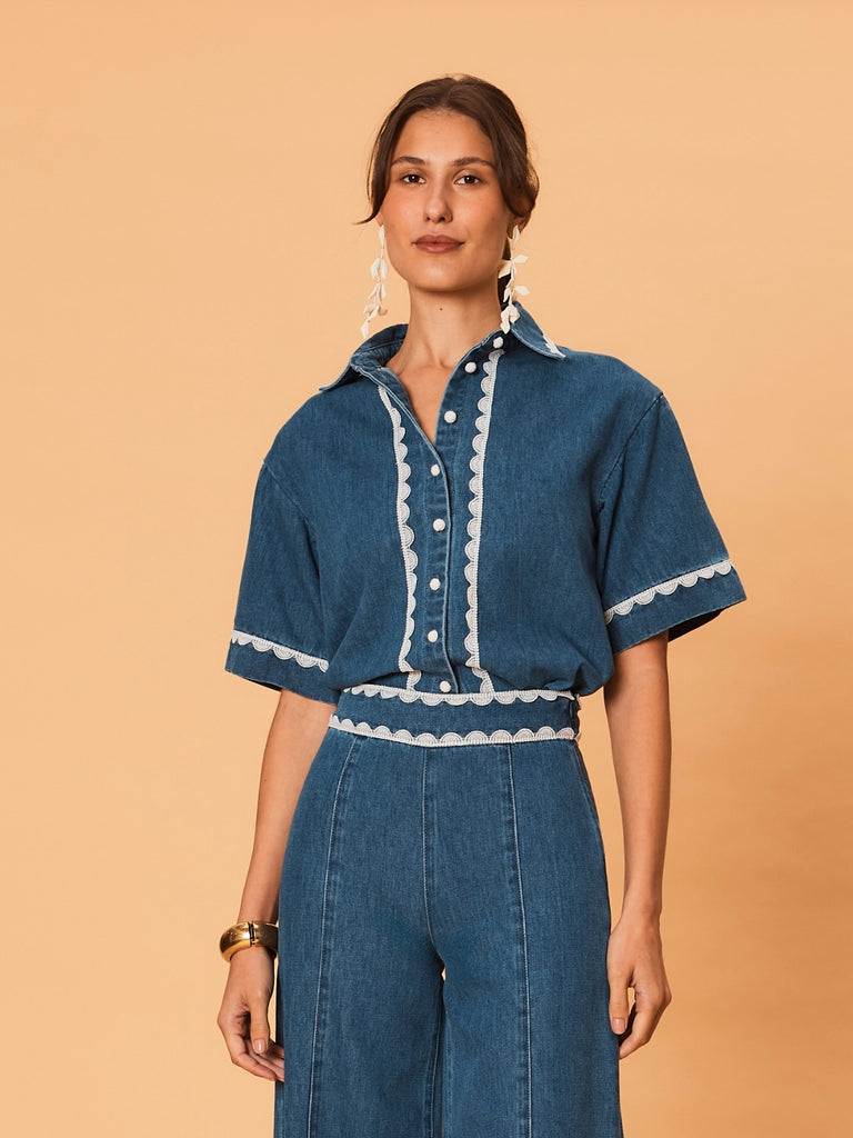 A young woman wearing a blue La Maise Ellie denim shirt with white lace trim on a beige background.