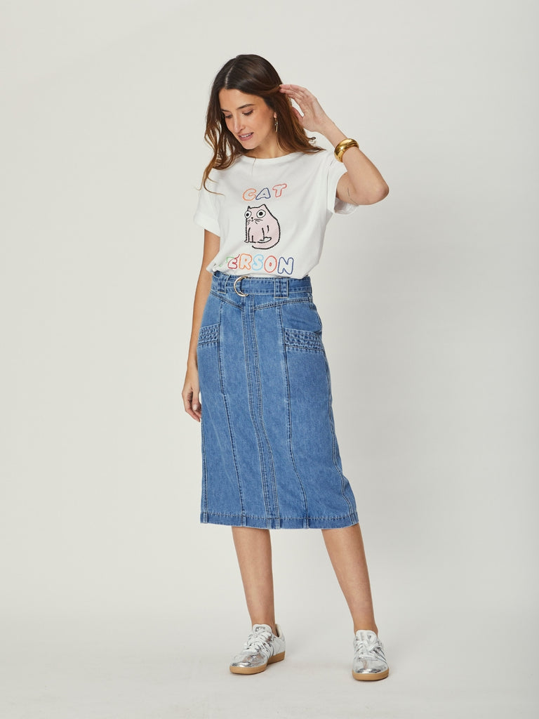 A woman wearing a white WATO tee with a graphic design and a blue denim skirt on a plain background.