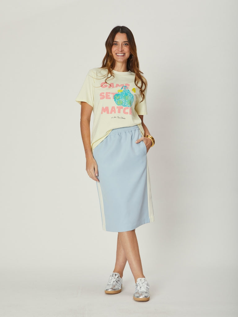 A woman wearing a graphic WATO tee and a light blue skirt on a plain background