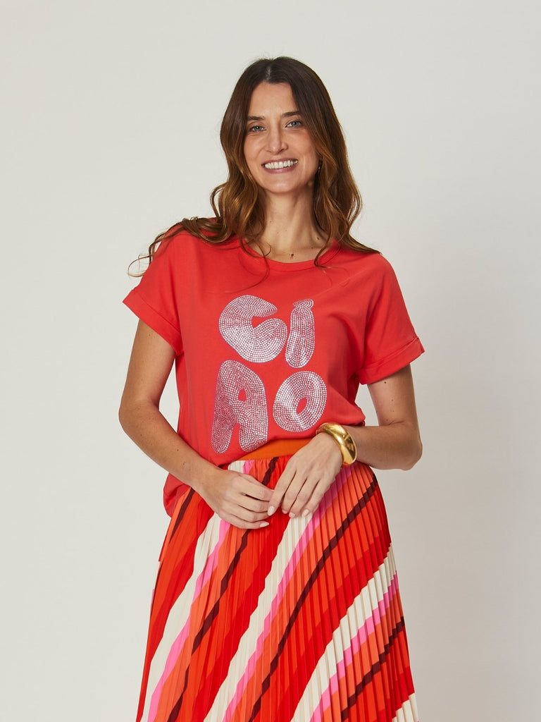 Woman wearing Jade Relaxed Tee – Ciao Orange, a relaxed orange graphic T-shirt with bold “Ciao” lettering, styled with a pleated skirt.