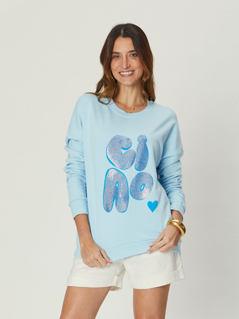 Woman wearing We Are The Others Amara Slouch Sweat – Ciao Sky Blue, a relaxed light blue sweater with a bold “Ciao” graphic, styled with white shorts.