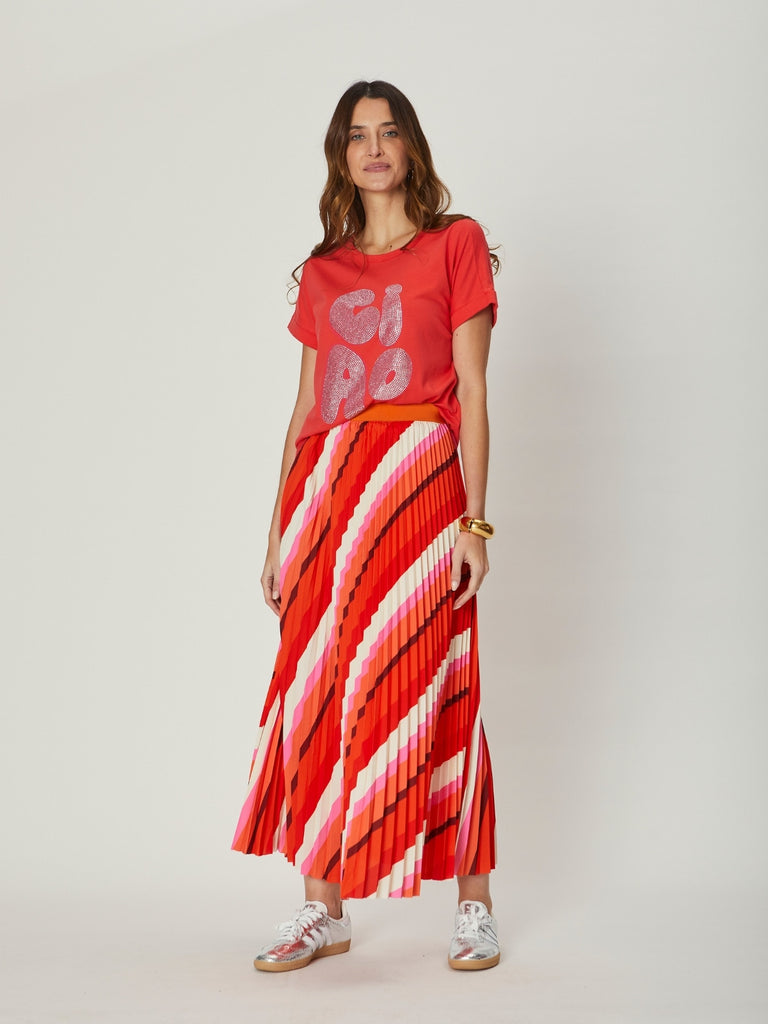 A woman wearing the Simone Pleat Skirt in red and pink stripes paired with a red graphic tee, showing the skirt’s flowing pleats and bold diagonal pattern.