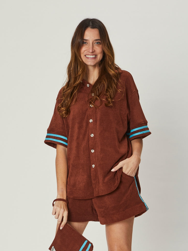 A woman wears the Stephanie Towelling Shirt in brown, a relaxed button-down with short sleeves and blue stripe detail, styled with matching shorts.