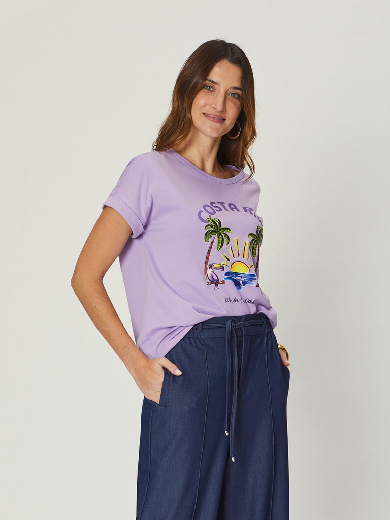 A woman with long brown hair smiles softly while posing in a lilac T-shirt and relaxed-fit indigo wide-leg pants with pockets and a drawstring waist.