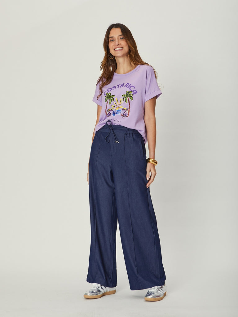 A woman with long wavy hair is standing and smiling, wearing a lilac graphic T-shirt tucked into wide-leg indigo pants with a drawstring waist, styled with casual sneakers.