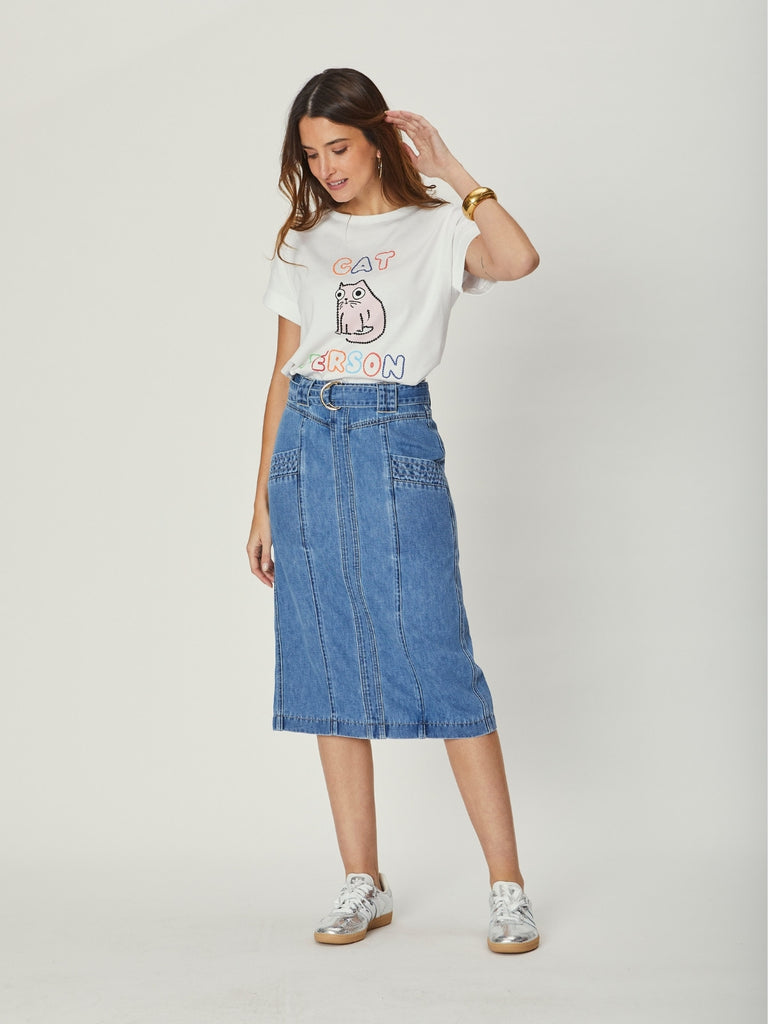 A woman wearing a white We Are The Other graphic tee and a high-waisted indigo denim midi skirt with pocket details and a D-ring belt, standing casually with hands at her sides.
