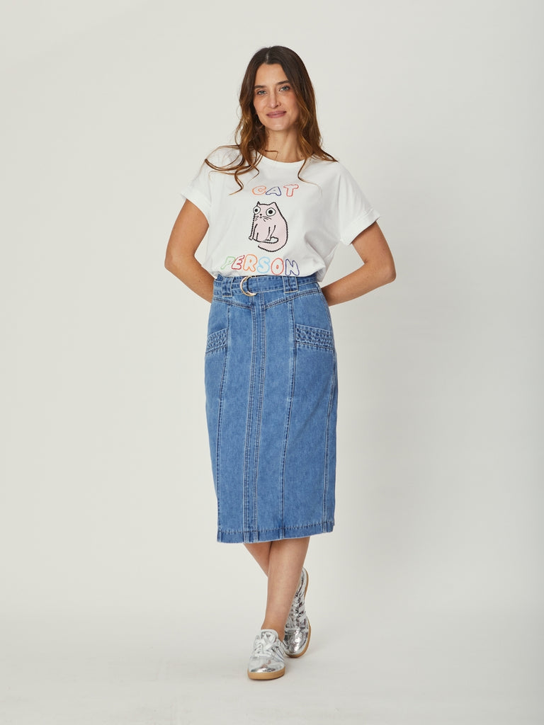 A woman wearing a white We Are The Other Cat Person tee and a high-waisted indigo denim Kimberly midi skirt with pocket details and a D-ring belt, standing casually with hands at her sides.