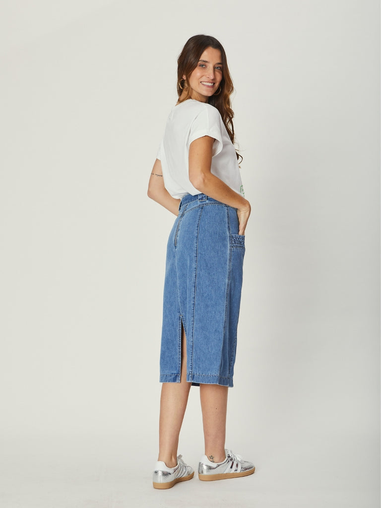 A back view of a woman wearing a white We Are The Other graphic tee and a high-waisted indigo denim midi skirt with pocket details and a D-ring belt, standing casually with hands in her pockets.