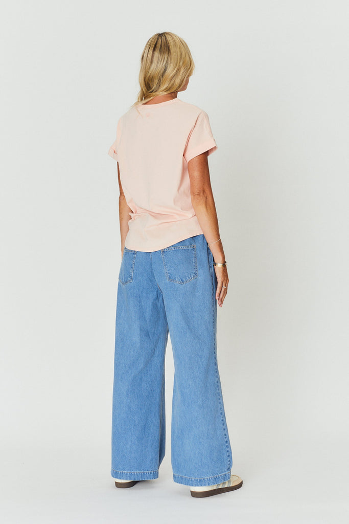 A woman wearing a light pink top and blue jeans standing against a white wall.