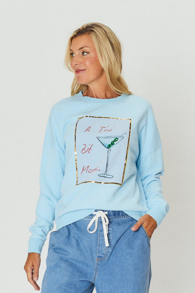 Woman wearing the Amara Slouch Sweat – Martini by We Are The Others, a light blue cotton sweatshirt with a martini glass graphic and gold foil border, styled casually with denim pants.