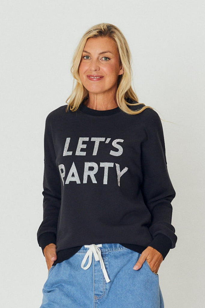 Woman wearing a black cotton sweater with silver slogan text reading "LET’S PARTY," styled casually with light denim pants.