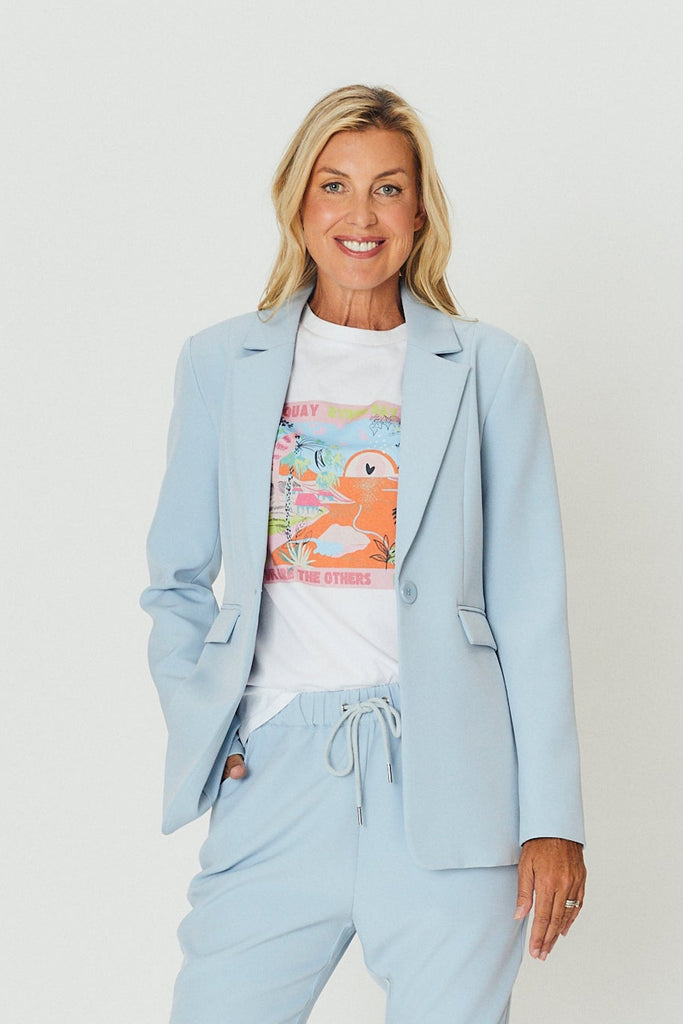 A woman wearing a light We Are The Others blue suit with a white shirt and sneakers on a white background.