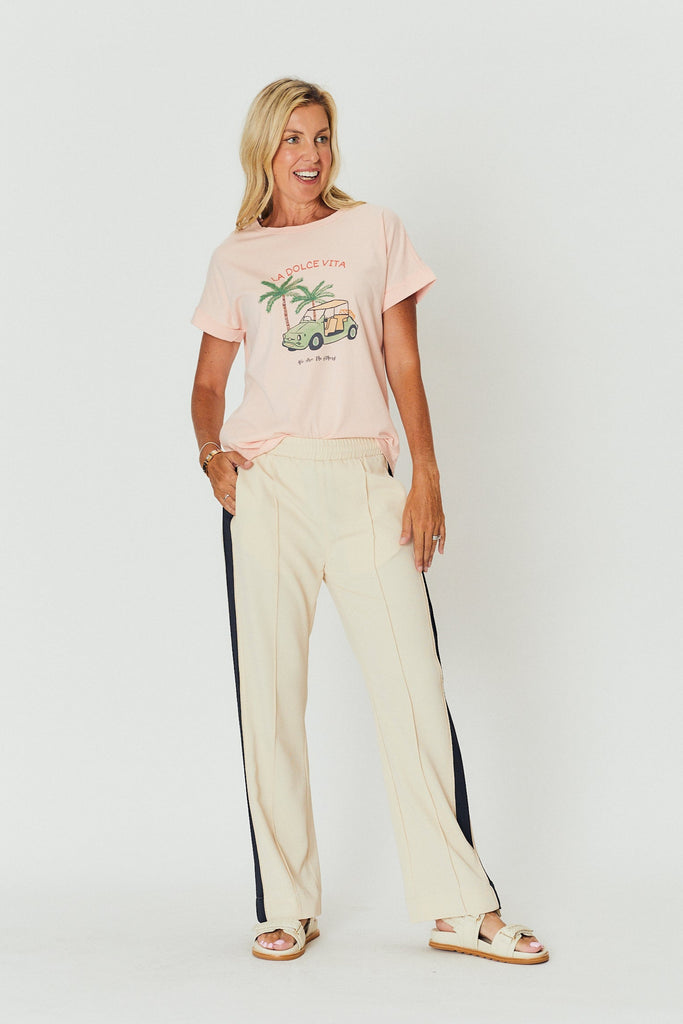 Woman wearing a We Are The Otehrs pink graphic Holiday t-shirt and beige Alba pants on a white background.