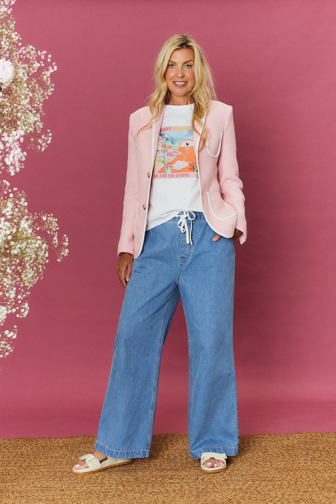 Woman in a pink blazer, white shirt, and blue jeans standing against a pink background with floral decorations.